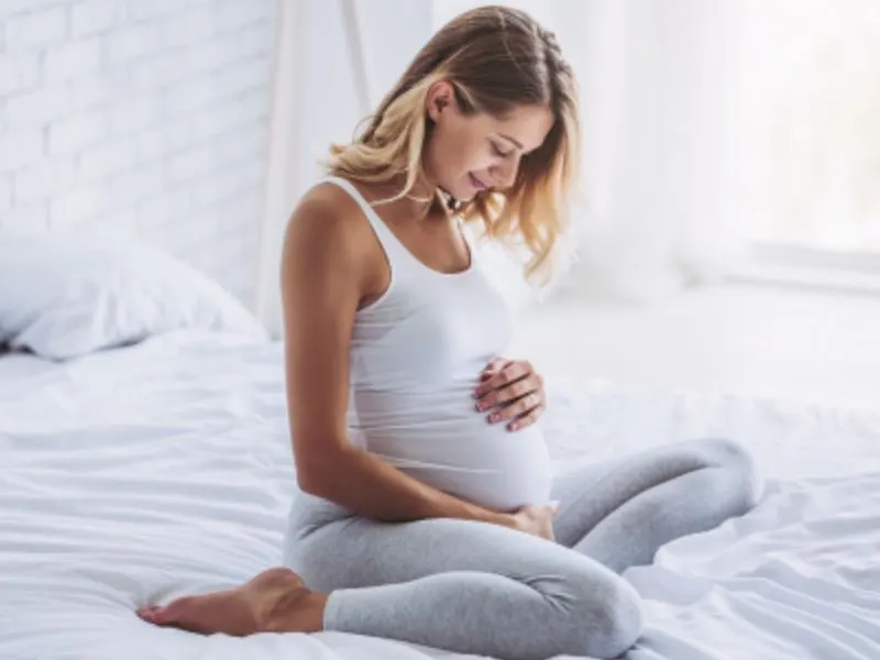 Pregnant woman sitting on a bed, cradling her belly with both hands.