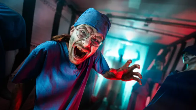 Person dressed as scary clown with blue hat and red nose reaching towards camera in coloured indoor lighting.