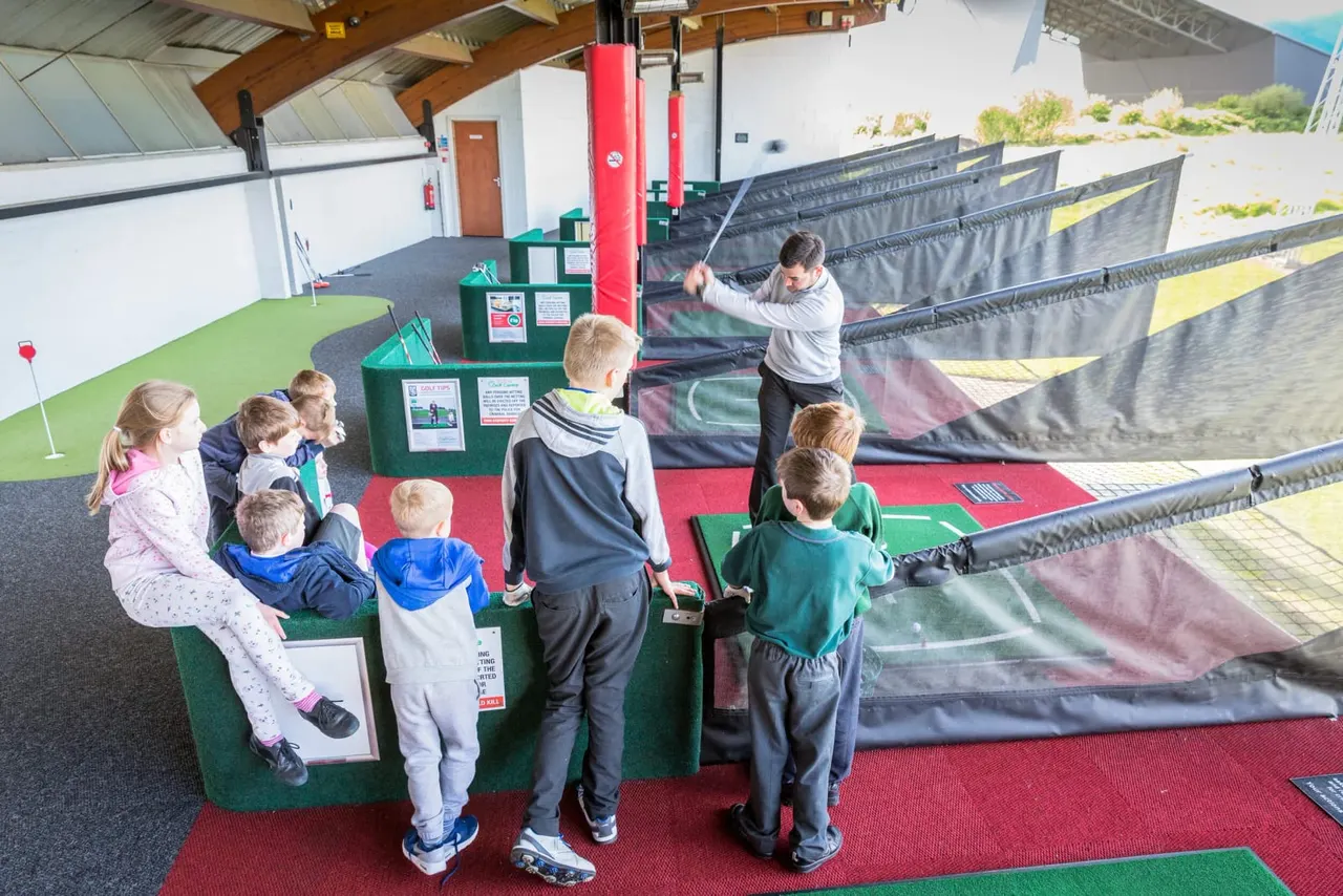 Children playing indoor mini golf with an instructor at a facility with colourful putting greens and netting.