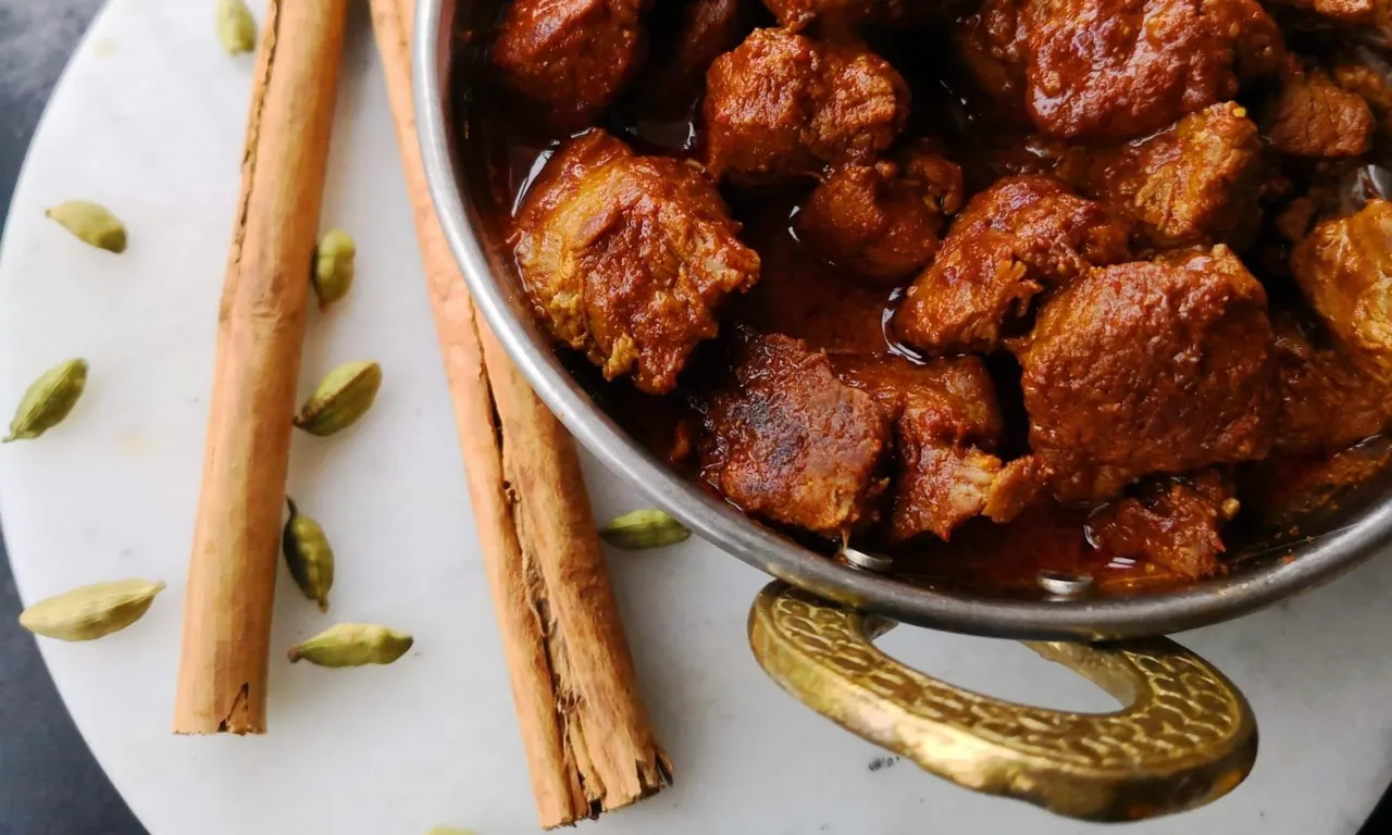 Bowl of spiced meat curry with cinnamon sticks and cardamom pods arranged on a white plate.