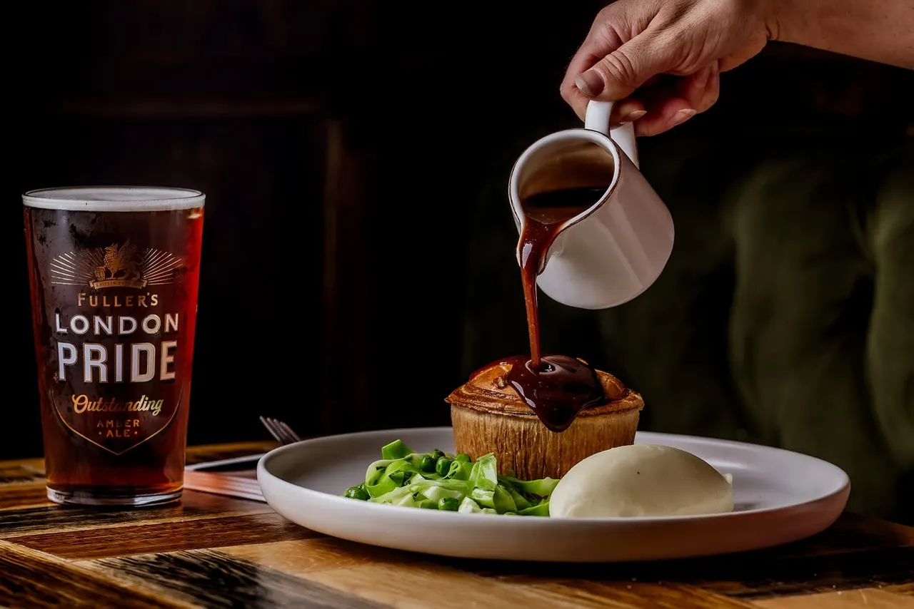 Hand pouring gravy over a pastry dish served with greens and mashed potato, alongside a pint of Fuller's London Pride beer.