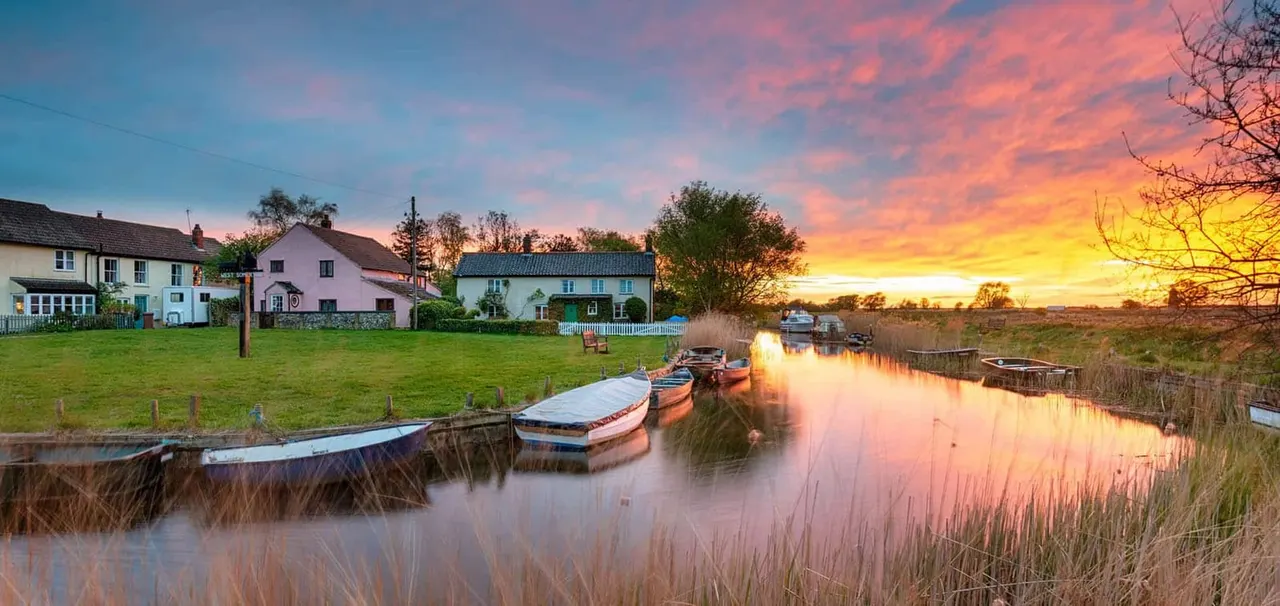 Moored boats on a calm waterway at sunset, with cottages and green fields beyond.