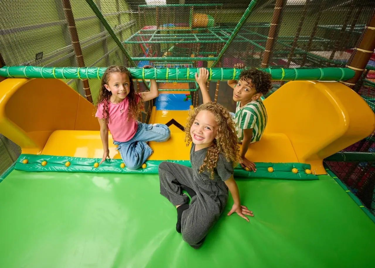Four children playing in an indoor soft play area with yellow slides and green netting.