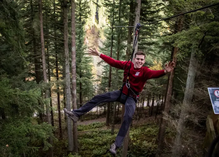 A young man on a zip wire between trees, arms outstretched, in a forest canopy course.