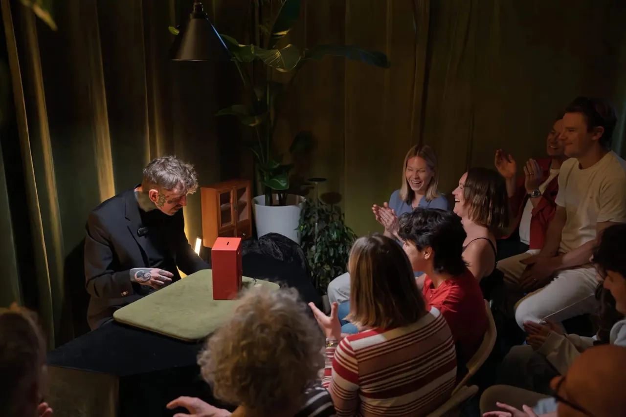 A magician in a dark suit performs a close-up magic trick with a red box to an engaged audience seated around him in an intimate venue.