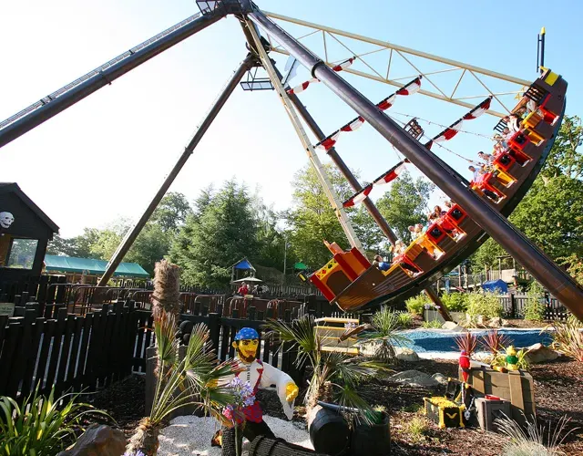 A red and yellow swing ride tilted dramatically at a theme park, with visitors and landscaped gardens visible below.