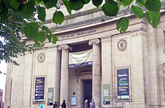 Classical stone building with columns and "Art Gallery Library & Museum" inscription, visitors entering the grand entrance with banners displayed.