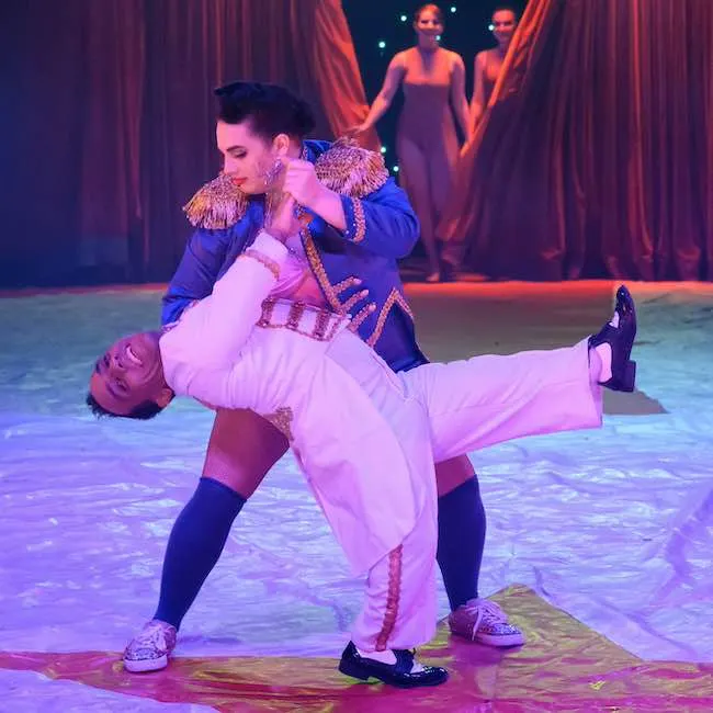 Circus performer in ornate blue jacket holds another performer in white outfit in a dramatic lift under red curtains and stage lighting.