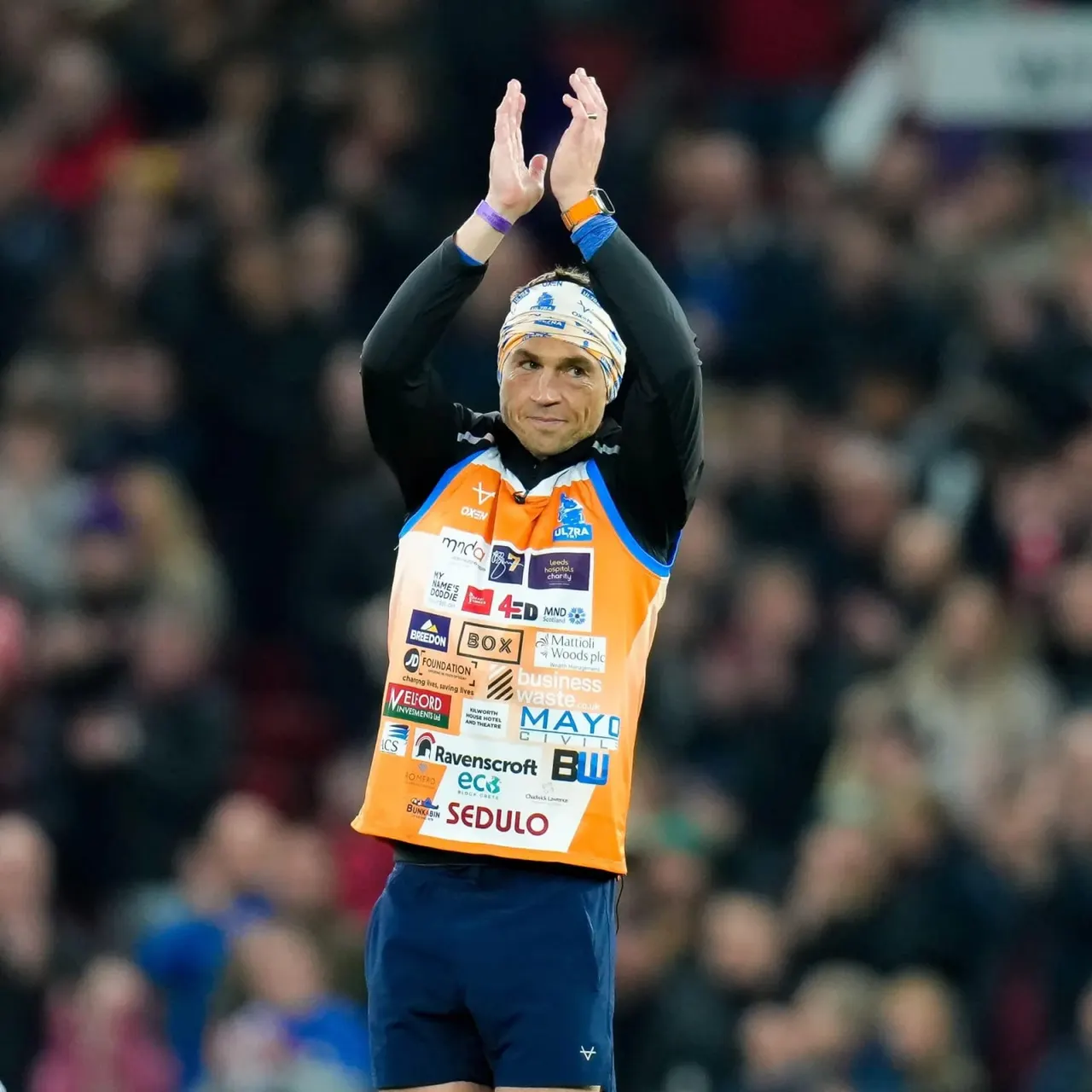 This is a photograph, not a designed graphic.

Athlete in orange and blue sponsor kit with headband raises both arms in celebration before a crowd.