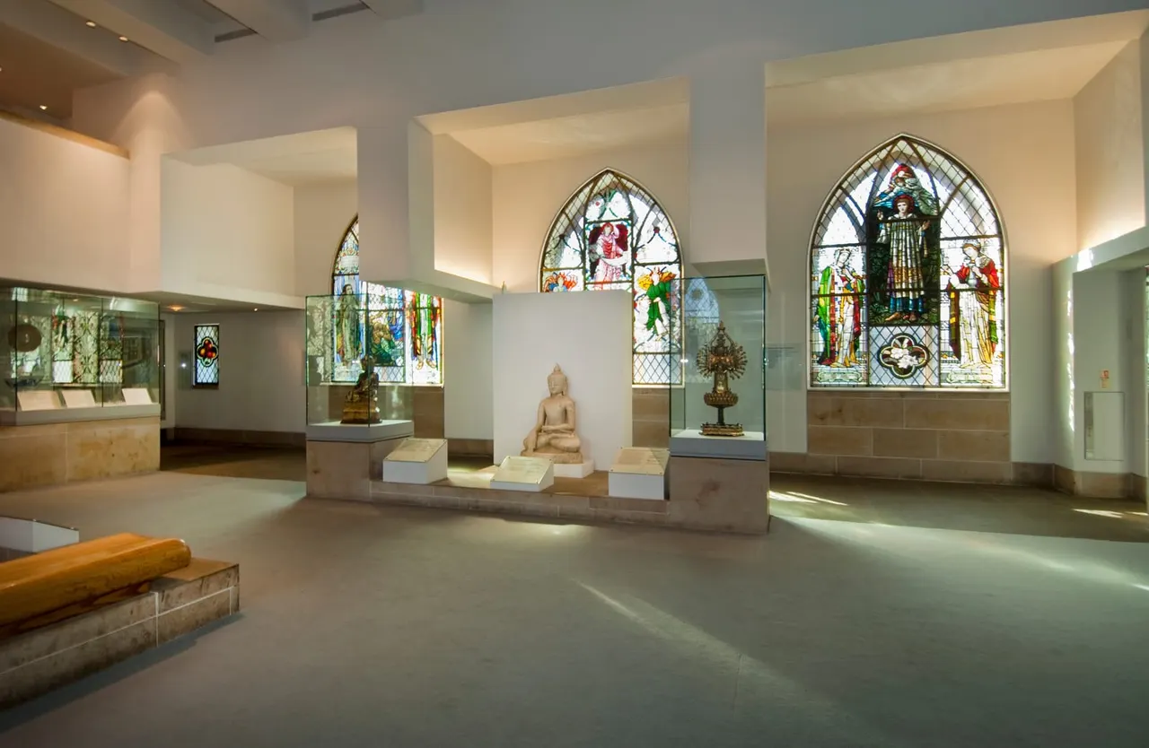 Museum gallery with stained glass windows, stone Buddha sculpture, and display cases beneath arched windows depicting religious figures.
