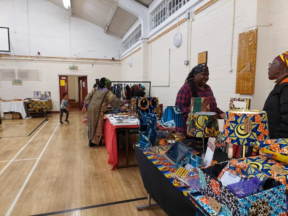 Indoor market stall with vendors selling colourful patterned textiles and crafts in a community hall.
