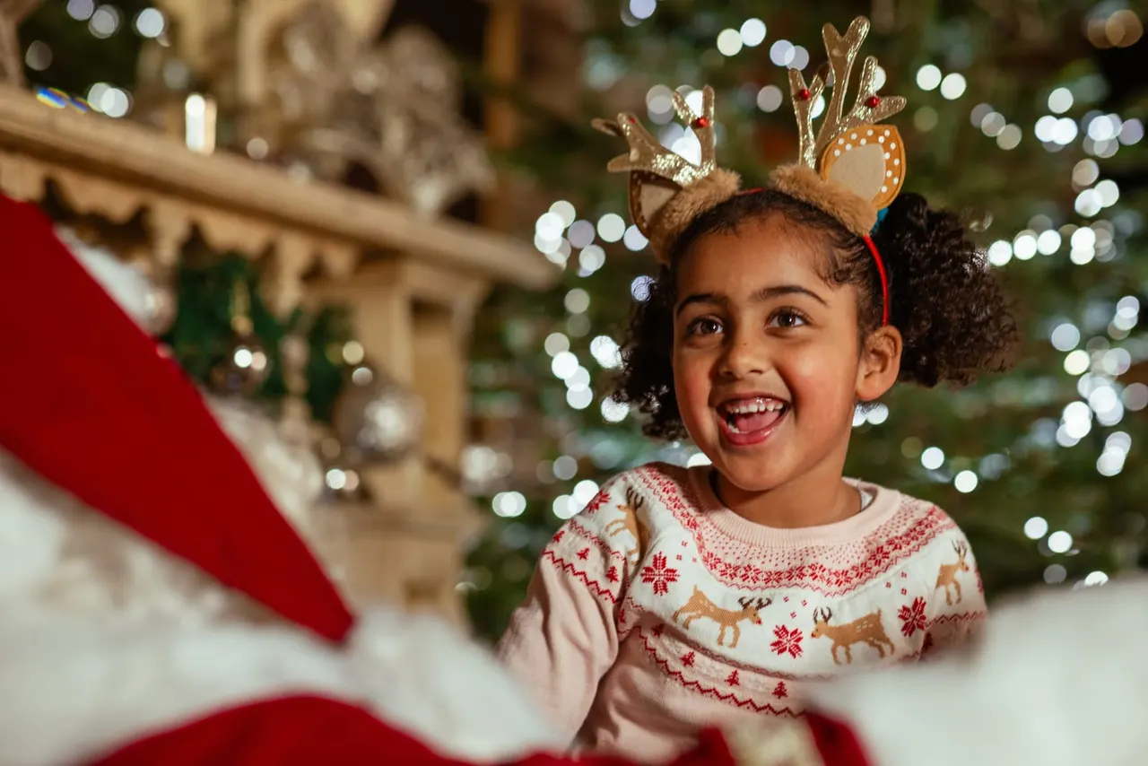 Child wearing reindeer antlers and festive jumper smiling in front of decorated Christmas tree and lights.