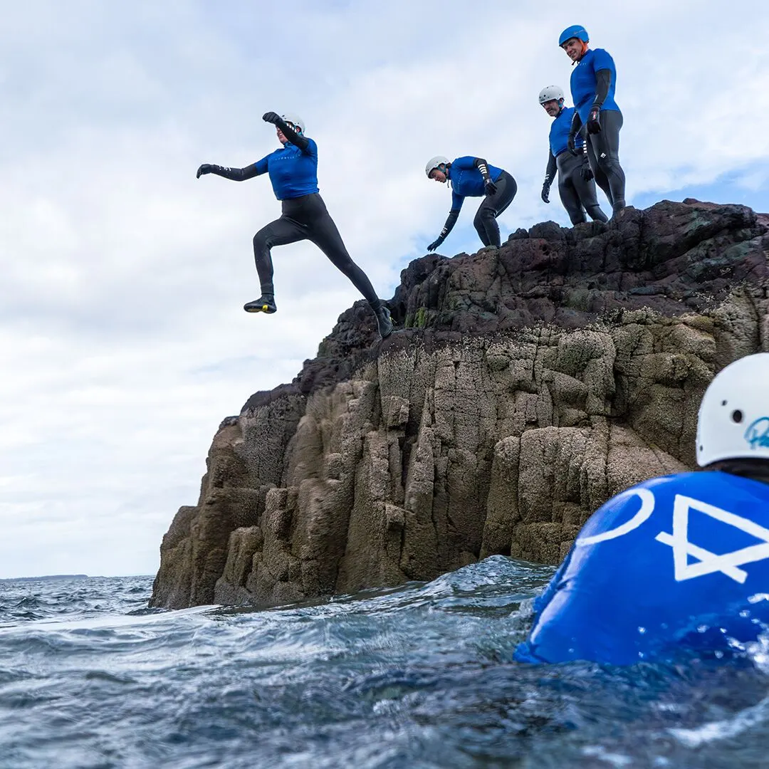 Person in blue wetsuit jumping from rocky cliff into sea whilst others watch from rocks above.