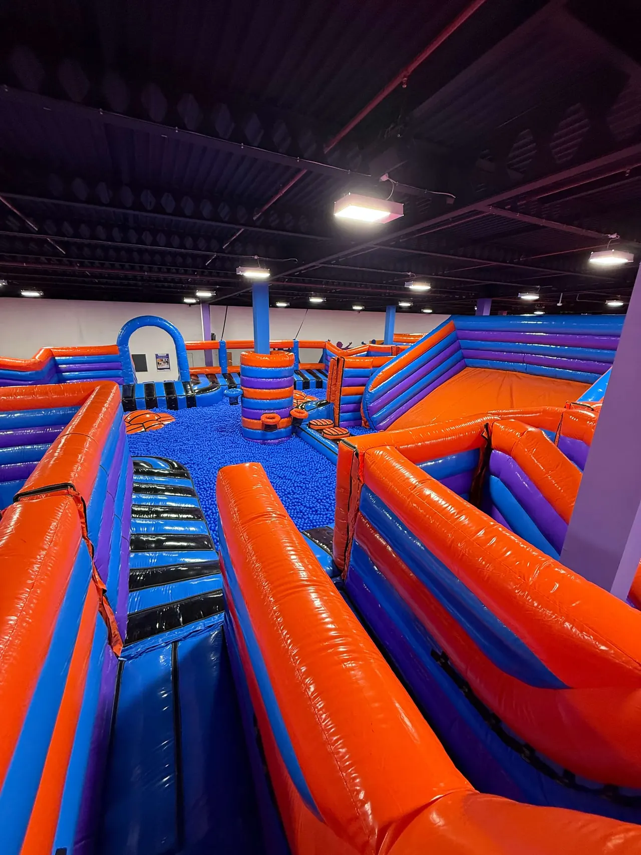 Indoor inflatable play arena with various bright orange and blue bouncy structures and obstacle courses.