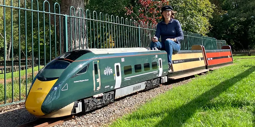 A person crouches beside a miniature passenger train with green and yellow livery on a track in a park setting.