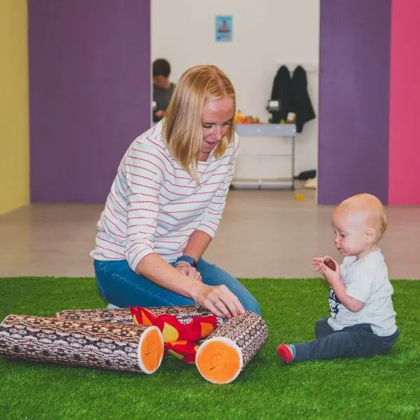 Woman and toddler playing with a toy log on artificial grass in a colourful indoor play space.