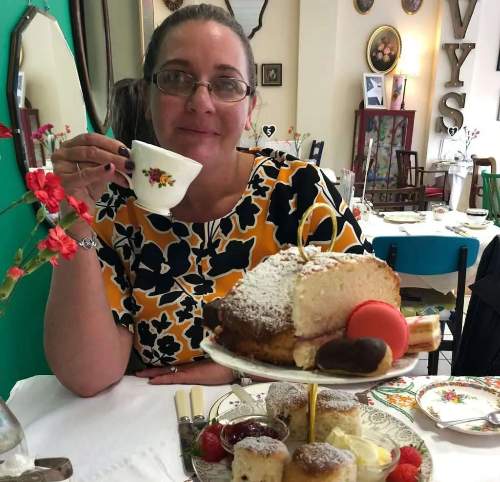 Woman enjoying tea and cakes at a decorated indoor café table with floral china and vintage décor.