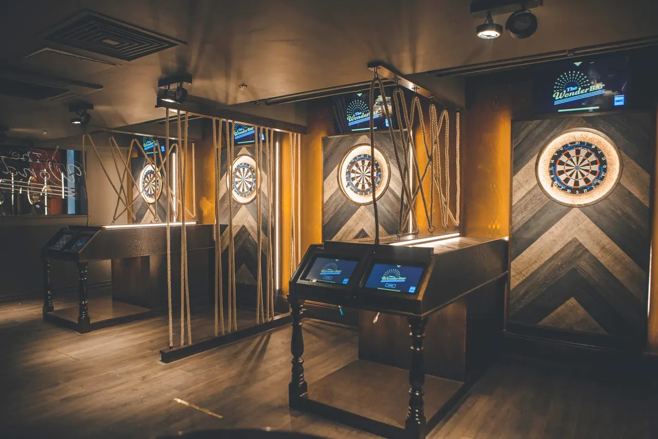 Indoor darts arena with multiple illuminated dartboards in enclosed booths, electronic scoring systems, and warm lighting throughout the venue.