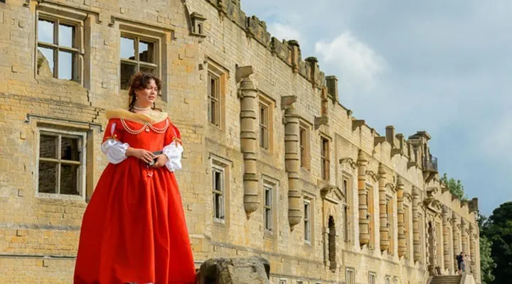 Person in red historical costume standing before stone castle building.