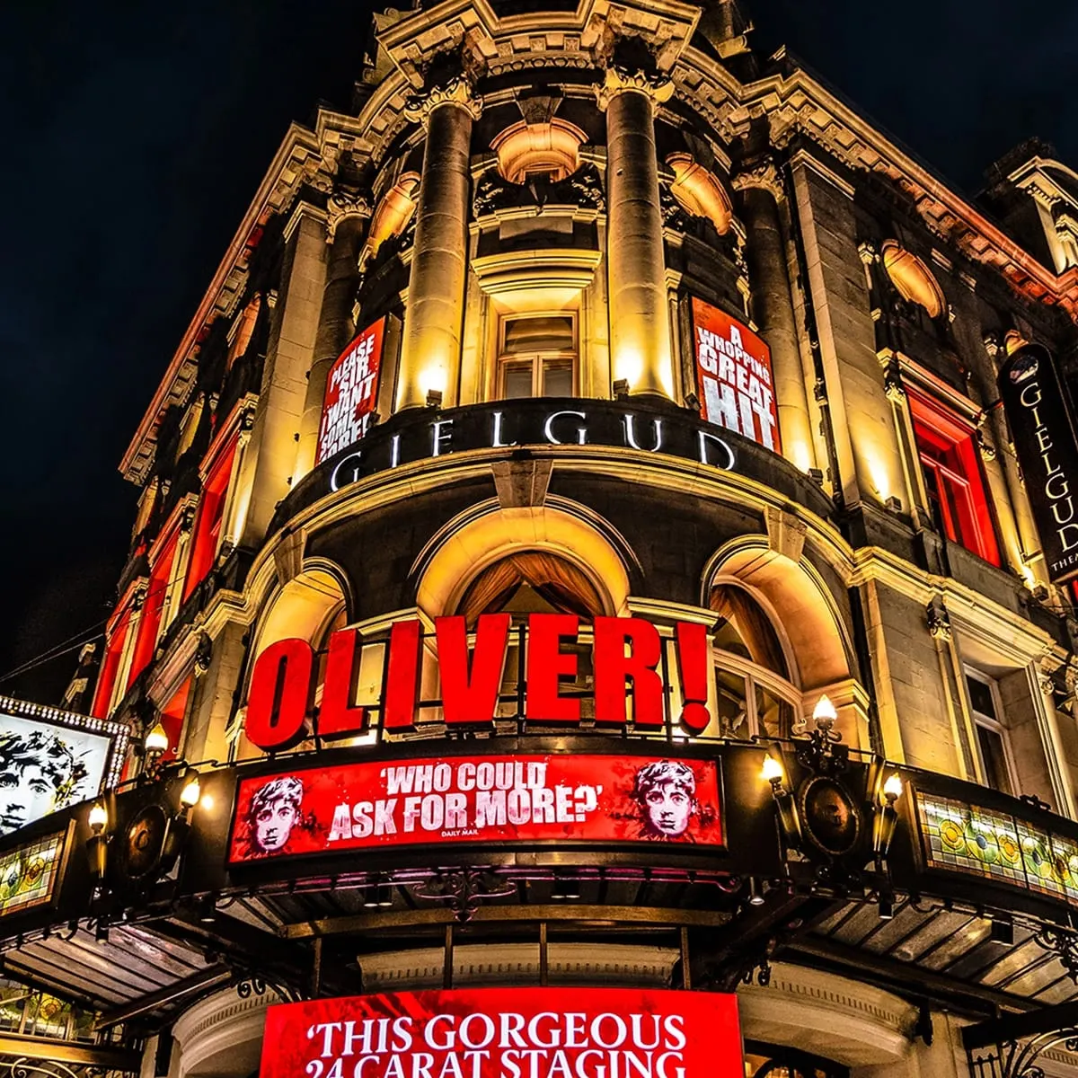 Night-time exterior of the Gielgud Theatre showing illuminated ornate Victorian architecture with red signage for the musical Oliver!.