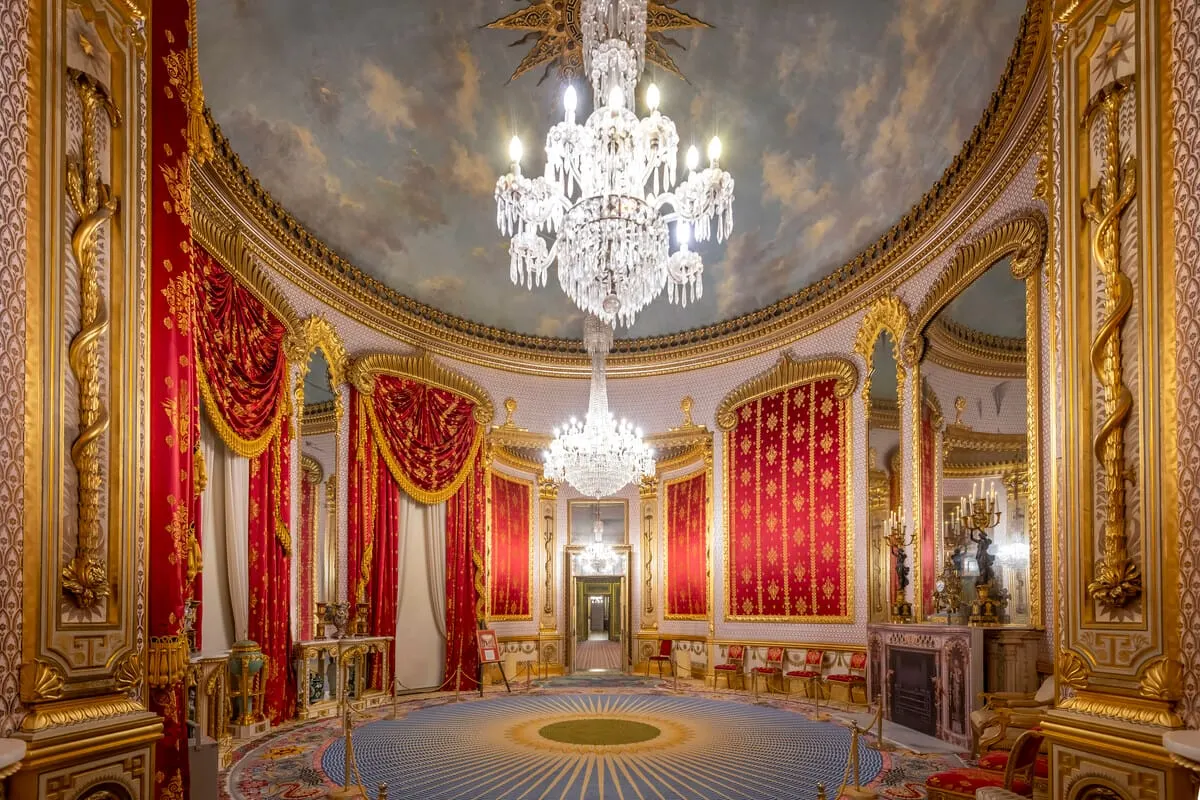 Ornate circular palace room with gilded walls, red velvet drapes, crystal chandelier and decorative painted ceiling.
