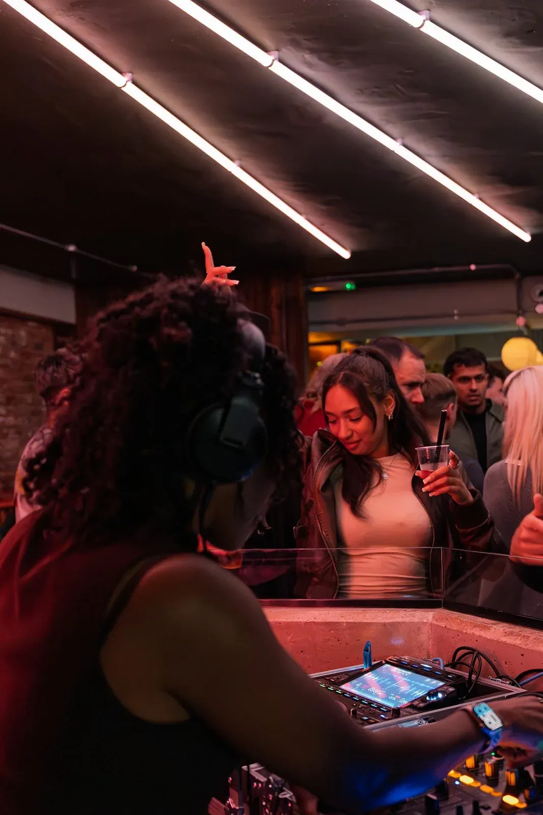 People socialising at a bar under red neon lighting, with a DJ booth visible in the foreground.