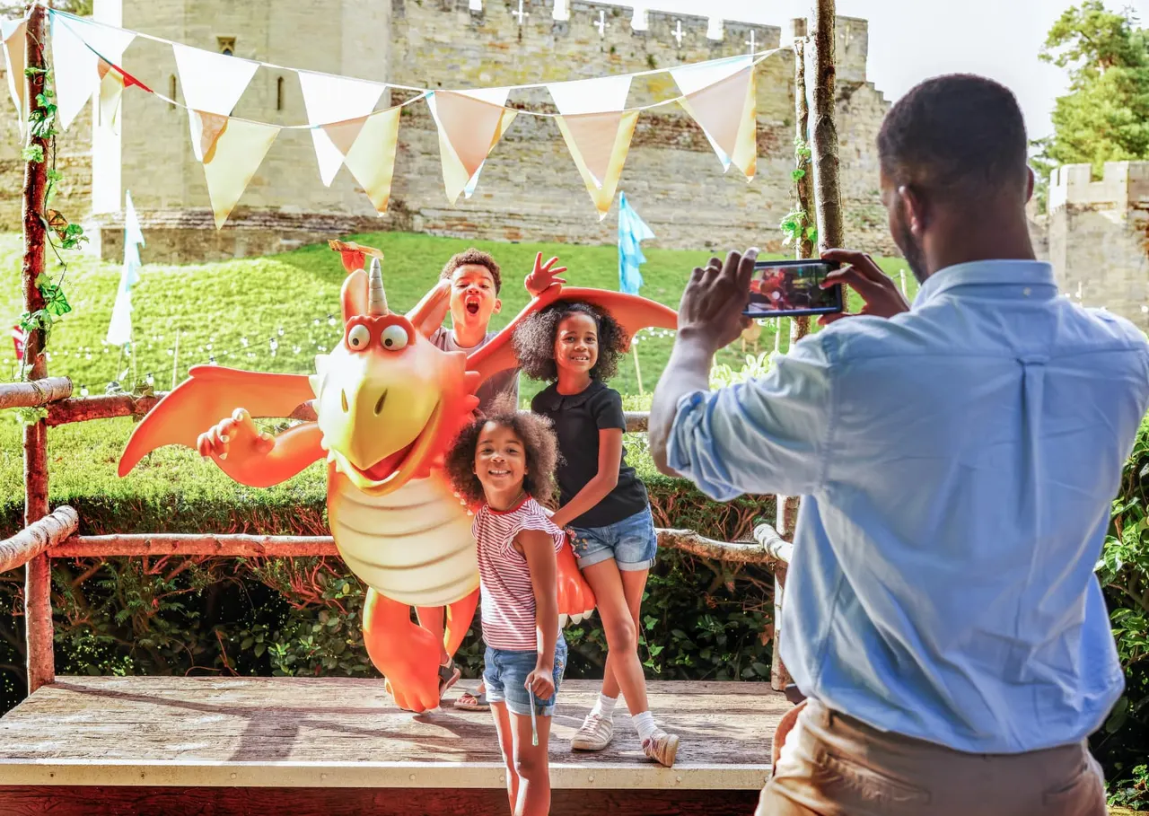 Man taking a photo of children posing with a large red and yellow dragon character at a castle venue with bunting and green lawns.