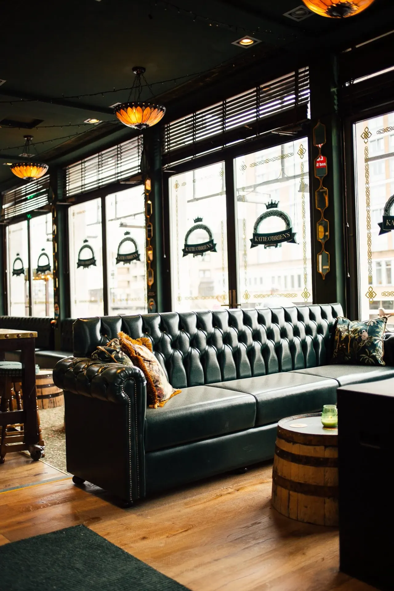 Upmarket bar interior with a dark green leather Chesterfield sofa, amber glass pendant lights, and large windows overlooking a street.