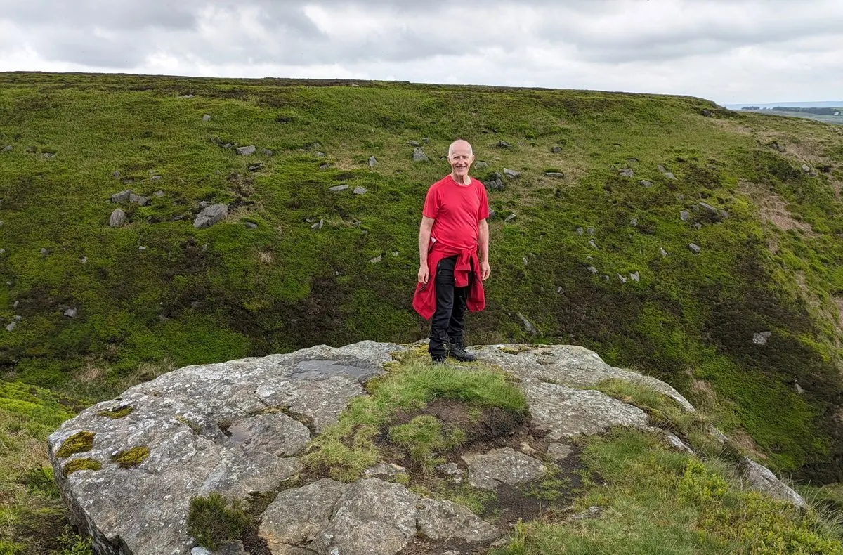 A person in a red shirt stands on a large rock outcrop overlooking moorland with scattered boulders and green grass.