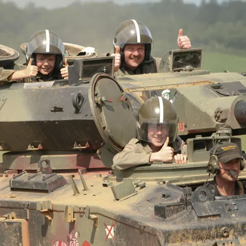 Three people wearing military helmets emerge from hatches in a vintage tracked military vehicle during a museum demonstration.