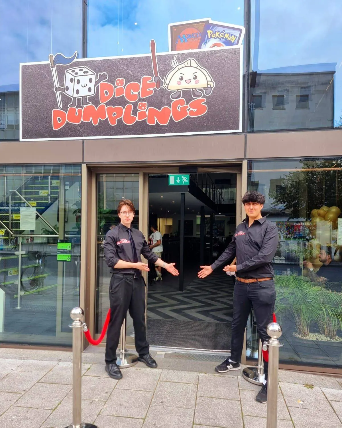 Two staff members in dark uniforms stand outside the entrance to Dice & Dumplings restaurant, welcoming guests.