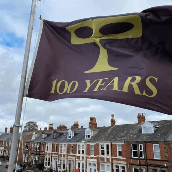 A brown flag with a yellow theatrical mask logo and "100 YEARS" text flies above a row of terraced Victorian buildings.