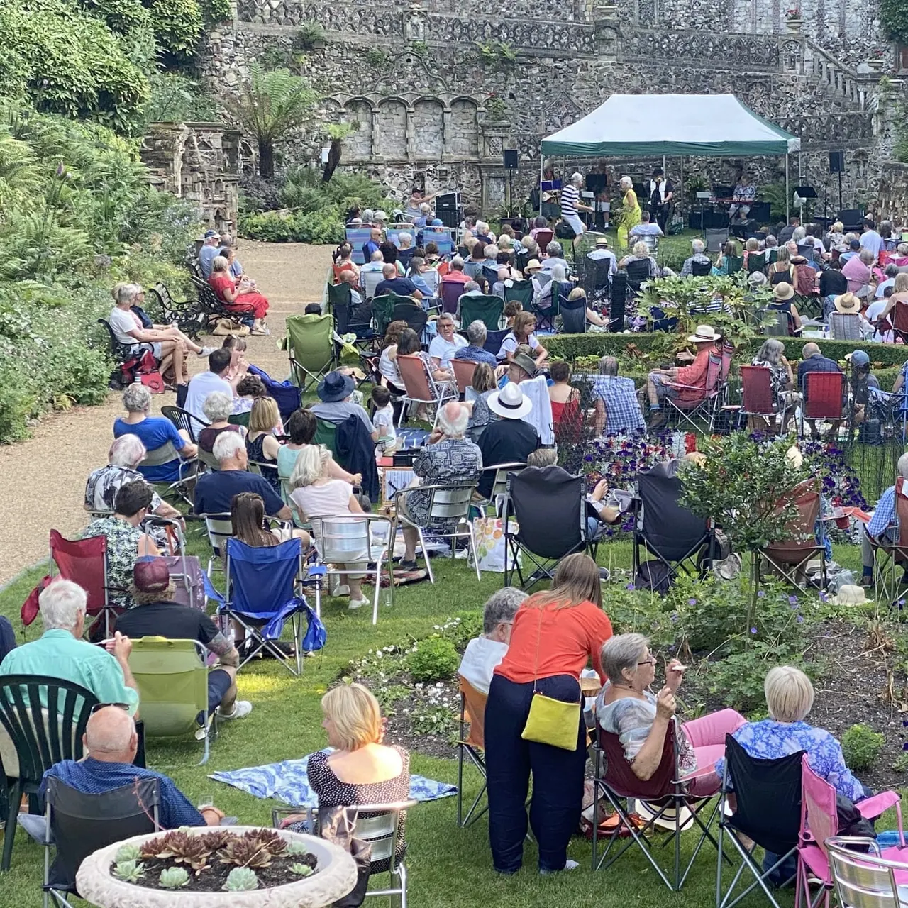Large outdoor event in a walled garden with many seated spectators facing a stage with green canopy, historic stone walls and lush greenery visible.