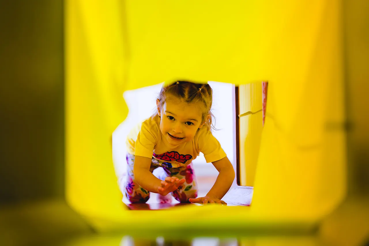 Smiling toddler in yellow shirt crawling through bright yellow play tunnel.