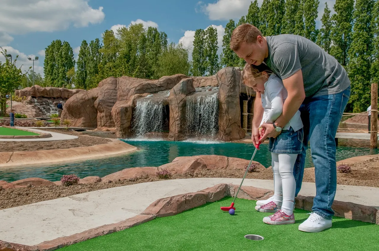 A man helps a young child play miniature golf on a bright sunny day, with a cascading waterfall and turquoise pool visible behind them.