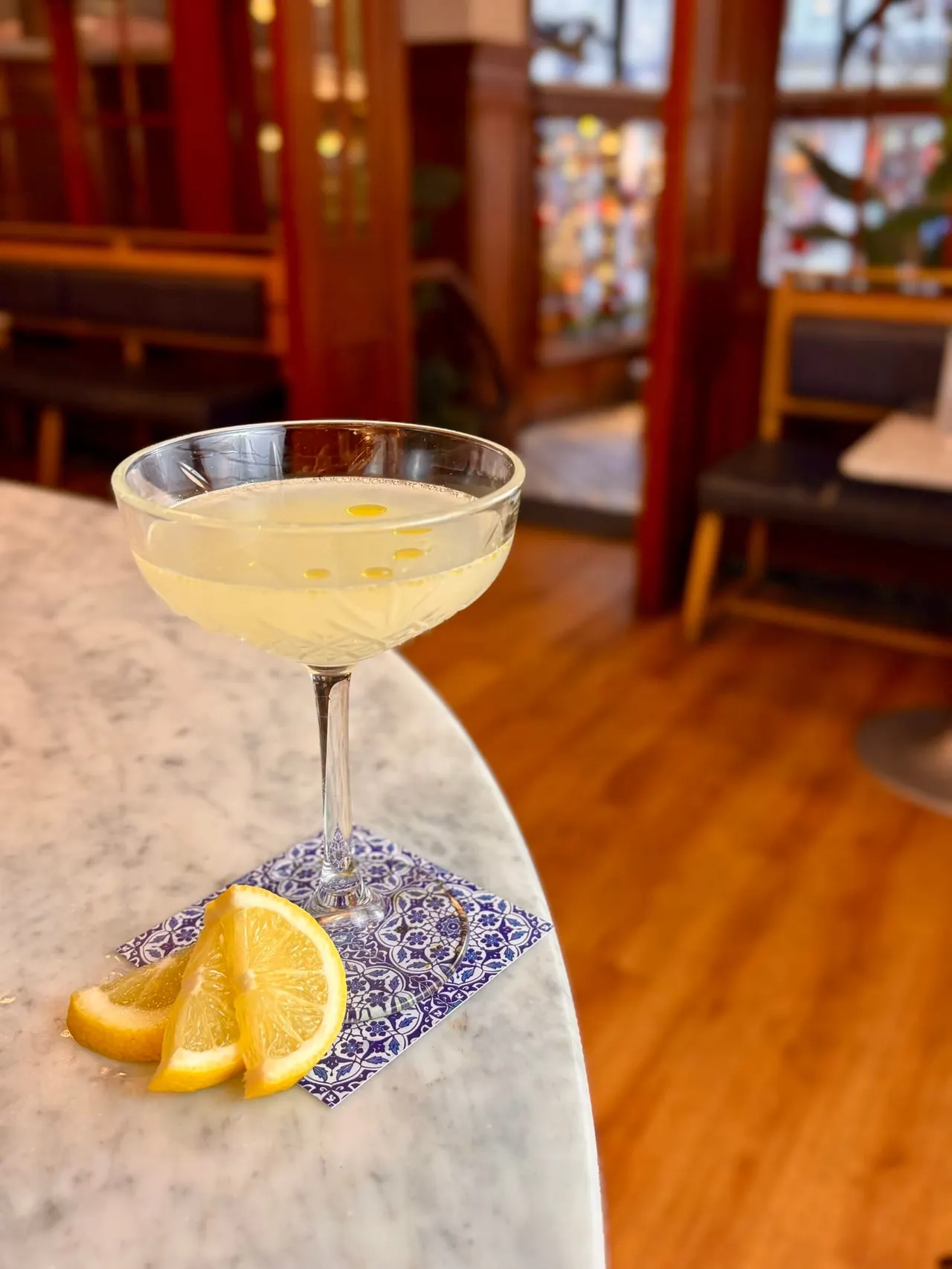 A pale yellow cocktail in a coupe glass with lemon slices sits on a marble table in an elegant red and gold interior.