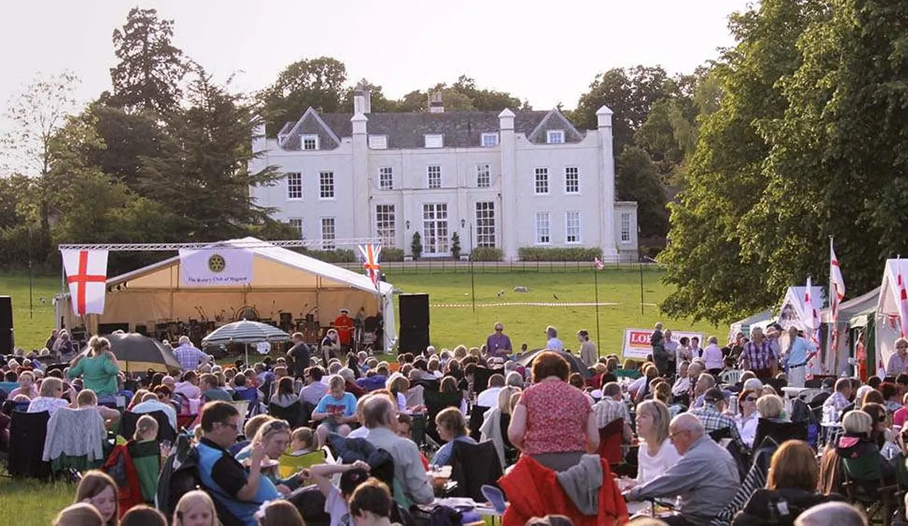 Large outdoor event with hundreds of visitors gathered on a lawn in front of a grand white mansion, with marquees, flags and vendor stalls.