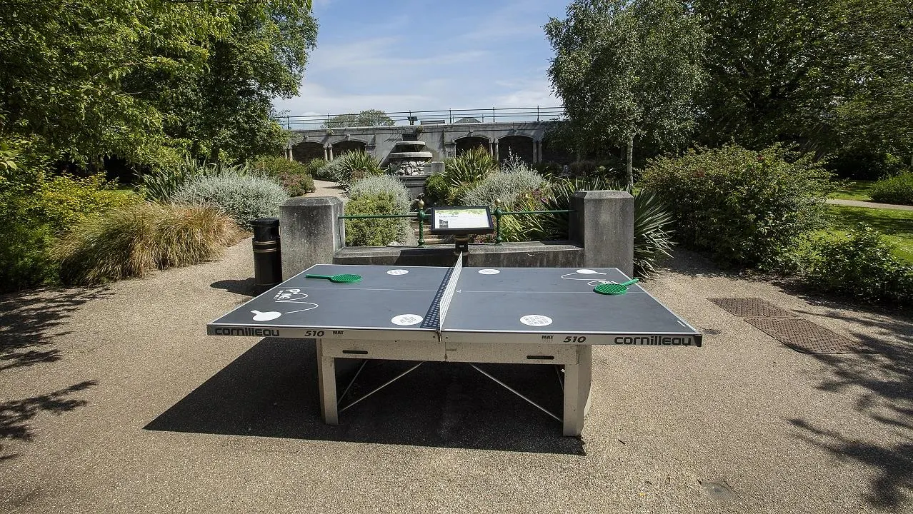 Table tennis tables set up outdoors in a leafy park area with a bridge and canal visible in the background.