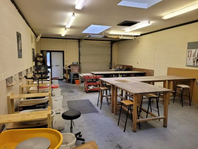 Interior of a pottery studio with wooden work tables, stools, and ceramic equipment including a kiln and potter's wheel.