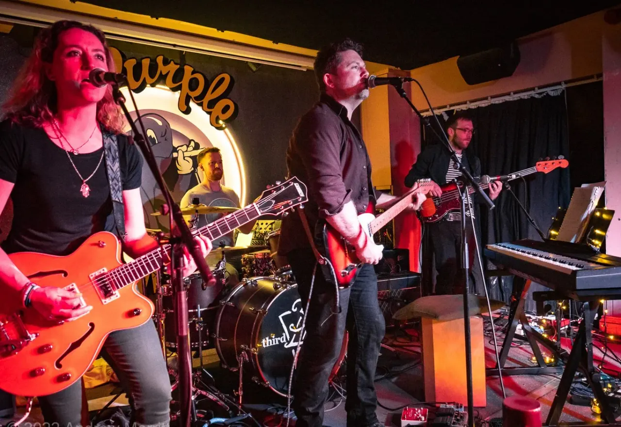 A live band performs on stage with a female vocalist and guitarist on the left, male singer in the centre, and guitarist and keyboardist on the right, lit by red and yellow stage lighting.