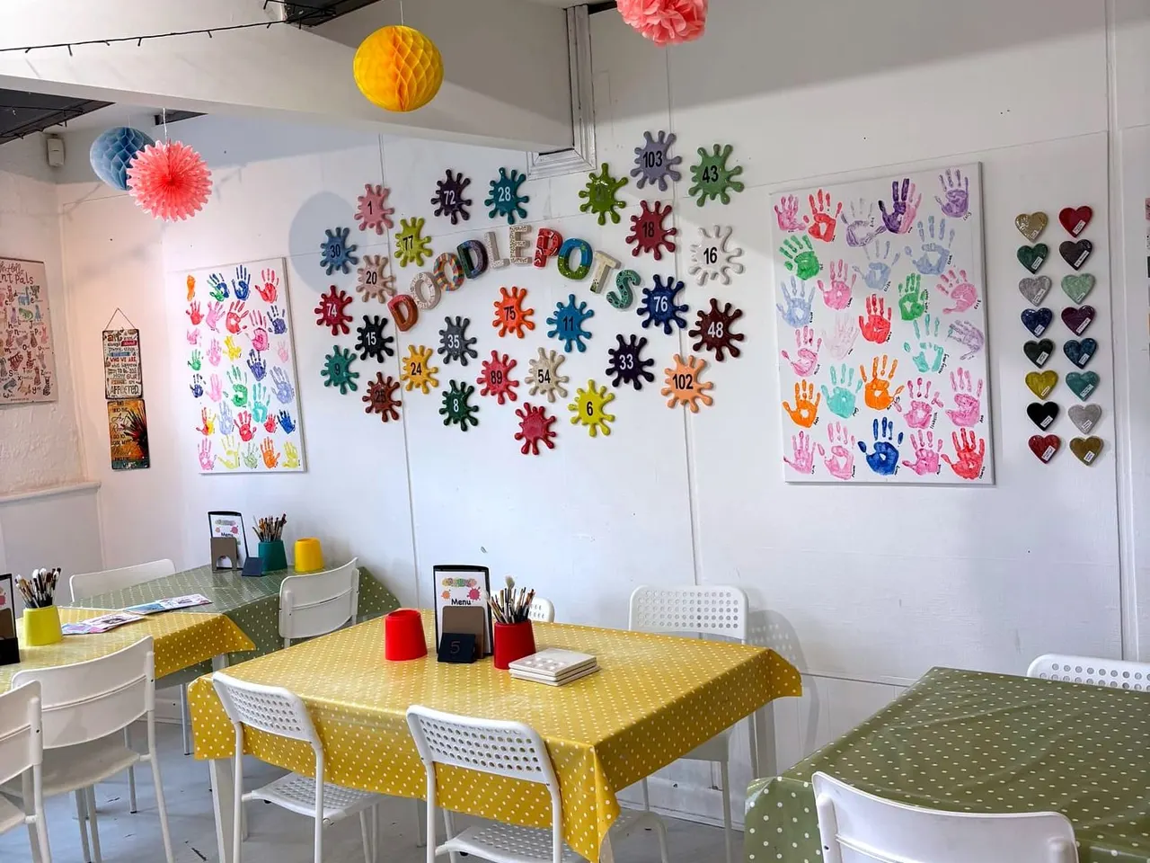 Interior of a children's craft studio with activity tables, colourful handprint artwork and decorative hanging ornaments on the walls.