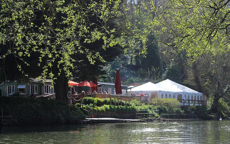 Riverside leisure venue with waterside café, red umbrellas, white marquee and diners beneath overhanging trees.