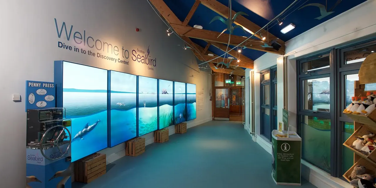 Interior of the Scottish Seabird Centre showing welcome signage, interactive displays with underwater marine life imagery, and exhibition spaces.