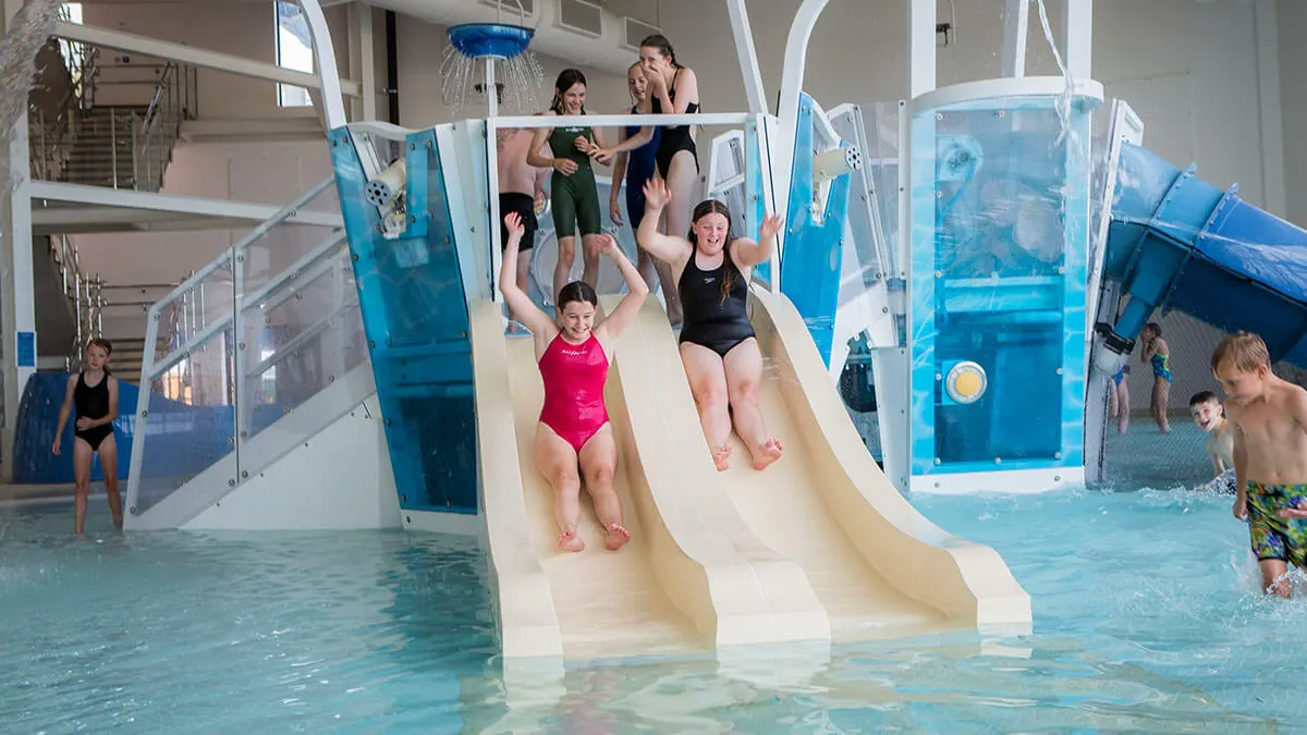 Children sliding down a water slide into an indoor swimming pool with supervisors and other visitors nearby.