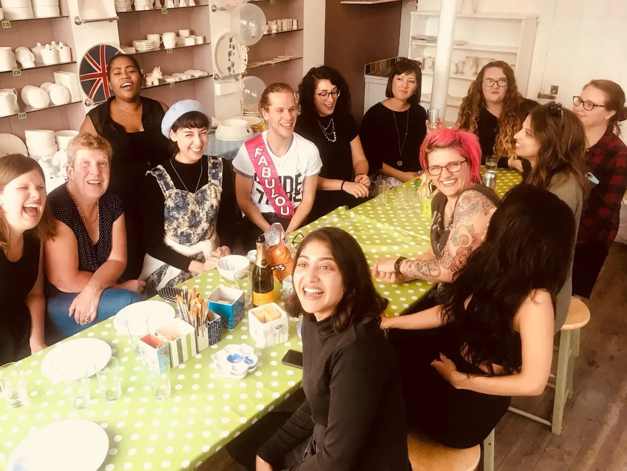 A group of smiling people gathered around a table set for a meal in a pottery studio with shelves of ceramics.