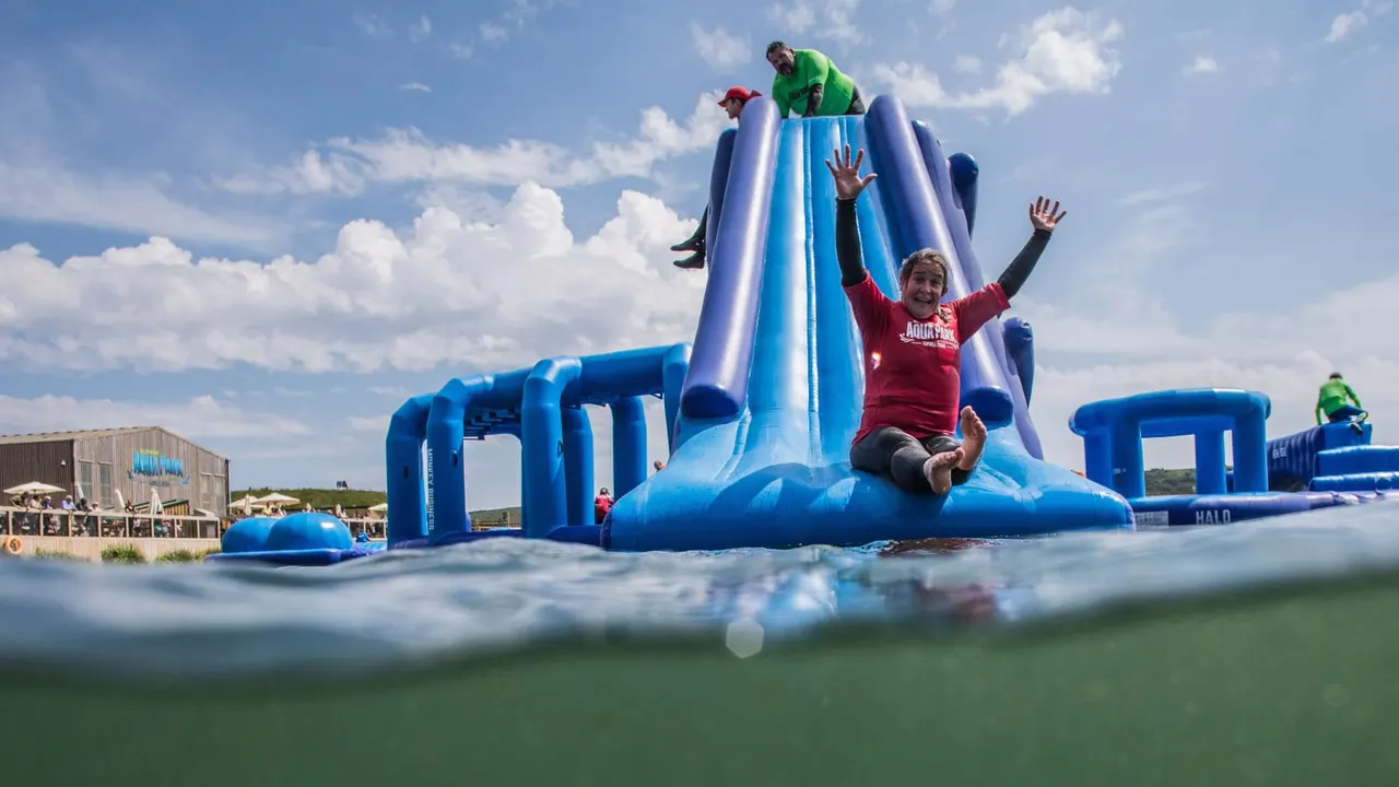 Child sliding down a large inflatable water slide whilst adults supervise on the blue obstacle course in shallow water.