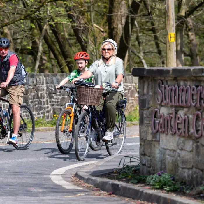 Three cyclists including a woman and child riding on a tree-lined path past a stone sign.