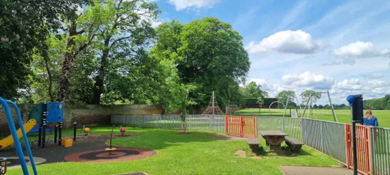 Children's play area with climbing structures, swings, slides and picnic tables set in a grassy field bordered by mature trees.