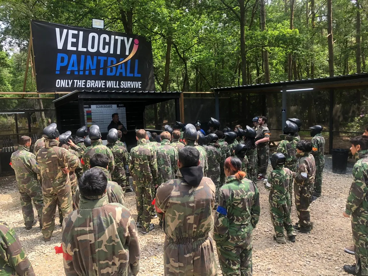 Large group of players in camouflage and protective gear gathered at a paintball venue briefing area under trees.