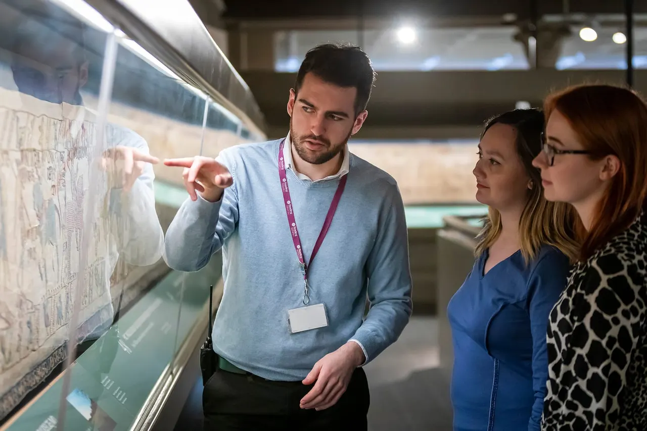 A male tour guide wearing a lanyard points to a display case whilst explaining it to two female visitors inside a museum gallery.